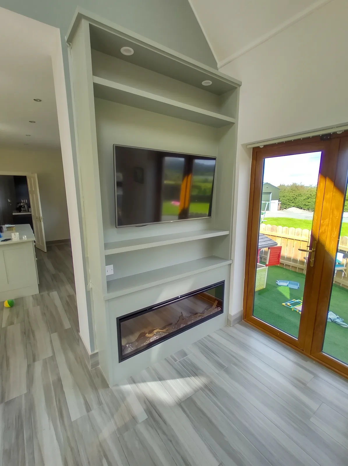 Sage-grey chimney-breast media unit with downlit shelving and a long inset fire in a vaulted room