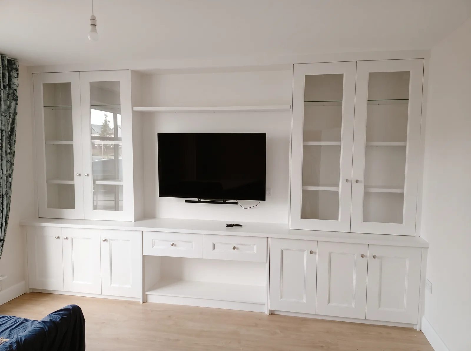 White wall of glazed display cabinets, Shaker cupboards and a central drawer unit beneath the TV